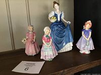 Four Royal Doulton figurines displayed on a wooden shelf: Helen (tallest) in blue dress holding fruit basket, Ivy in purple dress, two additional female figurines in pink dresses.