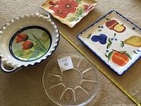 Four serving wares: a round floral ceramic plate, two square ceramic plates (one floral, one fruit-themed), and a clear glass serving bowl with handles.