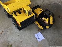 Yellow and black metal Tonka dump truck and scraper seen side by side on concrete floor. Both show wear.