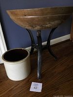Wooden bowl on three-legged metal stand next to tan crock on wooden floor