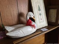 Side view of white carved wooden duck showing red ribbon and decorative details against wood panel background with framed wall art behind.