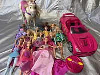 Photo of 11 Barbie dolls in various outfits, a white Barbie horse with pink bridle, a large pink Barbie convertible car, and a small pink Barbie remote control car.