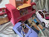 Pink and peach Barbie Dream Camper fold-out toy vehicle with kitchen section, purple and white convertible toy cars, and four dolls (three female, one male) laid in front.