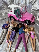 Pink Barbie Jeep with white roof and teal seats next to six Barbie dolls and one Ken doll lying on the ground