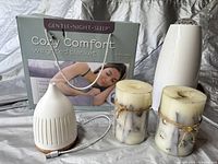 Photo showing Cozy Comfort 10lb weighted blanket in box with woman sleeping on front, white air purifier device, white diffuser, and two pillar candles all displayed on silver sheet.