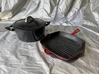 Photo of cast iron cookware set showing black Dutch oven with lid and red square grill pan with handle on a fabric surface.
