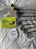 Golf clubs arranged on a white sheet with boxed golf balls, wooden tees, and can sleeves. Clear view of club heads and packaging.