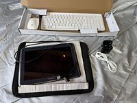 Samsung tablet with connected USB cable, white keyboard in box, white mouse, black charger, white and black fabric sleeves.