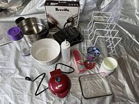 Assorted kitchen items including Breville Smoking Gun in original box, metal and ceramic bowls, squeeze bottle, kitchen timer, measuring cup, plastic cup, black rotary tool, oven mitt and white wire rack placed on fabric.