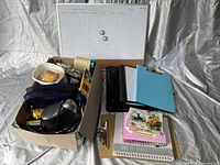 Photo showing the full box of assorted supplies and some binders and clipboards arranged beside it, along with a desk calendar pad in the background.