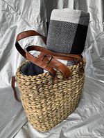 Front view of woven picnic bag with leather shoulder straps and checkered picnic blanket roll inside