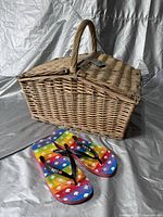 Closed wicker picnic basket with handle and a pair of rainbow-patterned flip-flops placed in front