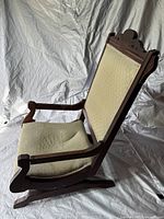 Side view of the antique wooden rocking chair showing the upholstered backrest and seat with wood carved armrests and rockers.