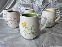 Six white ceramic mugs with different colored interiors and spring-themed designs arranged on fabric background