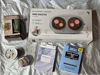 Lot overview with Sharper Image Mini Shiatsu massager box, hair serum container, wellness cards, Buddha scented diffuser, and additional candles