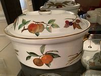 Wide view of white ceramic covered baker featuring painted peach fruit motif and gold trim, showing lid and body.