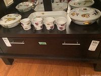 Lot displayed on dark wood furniture, showing two large covered bakers, eight mini bakers with lids, one rectangular tray with fruit and vegetable decorations and gold accents.