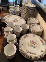 Wide view of stacked dinner plates, cups, saucers, and platters with floral pattern and gold rim.