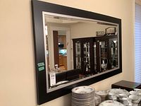 Front angled view of the rectangular mirror showing the black outer frame and silver inner border with beveled edges reflecting a room interior.