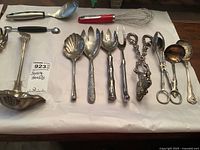 Overview of various serving utensils including ladles, a whisk, forks, tongs, scoops, and spoons with metal handles and an ornate tongs piece.