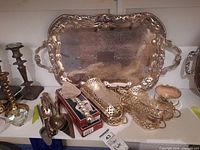 Photo showing the ornate rectangular silver plated serving tray with two handles, along with pierced pattern holders, cutlery and a small cup, arranged on a shelf.