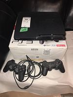 Sony PlayStation 3 console with 2 wired controllers placed on top of the original box labeled 120GB capacity. Console has dust on surface but no visible major damage. Controllers have wires attached.