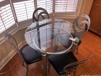 Full view of round glass top kitchen table with four metal frame chairs with black vinyl seats.