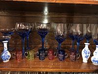 Front view showing six blue-stemmed wine glasses, two smaller blue-stemmed coupe glasses, four small colored glass mugs in red, green and pink, and two small white ceramic vases with blue floral decoration.