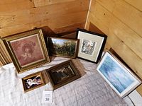Group photo showing six framed artworks arranged on a fabric-covered surface against a wood-paneled background, including varied sizes and styles such as portraits, landscapes, and urban scenes.