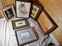 Six framed artworks laid on a cloth on the floor showing varied subjects: girl with dog, horse sketches, wood etchings, painting on feather.