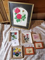 Full view of seven art pieces displayed on a cloth surface showing various floral prints and plaques, with one prominent framed rose print.