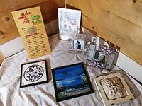Full view of seven-piece lot showing mixed wall hangings, paintings, prints, picture frames, and a decorative tile laid out on a cloth.