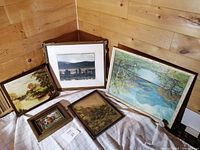 Five framed art pieces including paintings and prints of landscapes arranged on a cloth against a wooden wall.
