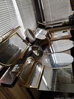 Photo showing various vintage and new mirrors including vanity, oval, square, and round shapes with mostly gold/brass and wood frames placed on a table near window blinds.