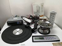 Mixed electronics and household items including a car radio, battery charger, thermostat, electric shaver, digital calculator, analog thermometer, and decorative metal bowl.