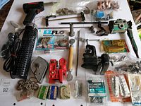 Photo showing assorted tools and hardware on a white surface, including a soldering gun, bicycle tire, clamps, bullet molds, and packaged items.
