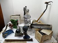 Photo showing multiple vintage tools including a metal spray canister, hand caulking tool with wooden handle, a large metal tube with bulb, a green seed spreader, and other assorted tools on a flat surface.