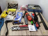 Photo showing a collection of mixed tools and accessories arranged on a wooden surface including clamps, screwdrivers, a measuring tape, and various drill bit packages.