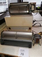 Photo showing the larger and smaller wood stove fans stacked on an environmental air system unit on a table, with window blinds in background.