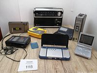 Overview of all vintage electronic items in the lot on a table including multiple cassette players, a portable speaker system, a Gameboy, and other devices.