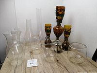 Lot showing three vintage glass oil lamps with decorative painted amber and multi-color bodies, surrounded by various clear and Dietz lantern glass chimneys on a wood surface.