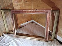 Two large wooden picture frames, one pink carved and one gold finish, shown overlapping and leaning against wood-paneled wall.