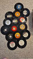 Photo showing an assortment of 45 RPM vinyl records laid out on carpet with visible various labels and some wear.