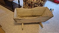 Shows exterior side view of antique wooden cradle with worn white paint and rocker legs