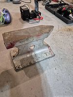 Side view of metal anvil showing flat top surface, horn, and base with mounting holes. Surface is worn and rusty.