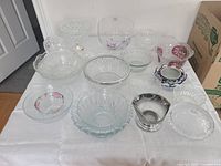 Photo showing a collection of 14 assorted glass and crystal bowls on a white sheet, various sizes and designs including clear, etched, patterned, colored and silver-tone pieces.
