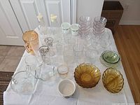 Photo showing a large assortment of glassware laid out on a white cloth. Includes pitchers, multiple glasses of different styles and sizes, amber bowls and dishes, and green small bowls.