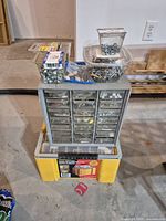 Front view of metal hardware organizer with multiple small transparent drawers filled with different screws, nuts, bolts, and washers. Various small plastic containers with additional hardware items placed on top of organizer cabinet. Yellow plastic storage box below organizer.