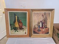 Two still life paintings framed in wood: one with vases and leaves, the other with bottle, glass and candle on table