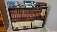 Overall view of wooden bookshelf filled with sets of encyclopedias, some volumes stacked on top.